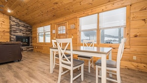 A log cabin interior with a fireplace, dining table, chairs, and large windows.