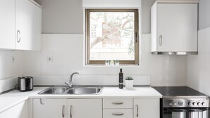 Una cocina moderna con armarios blancos, un lavamanos de acero inoxidable, un horno empotrado y un ventanal con vistas a un árbol y un edificio.