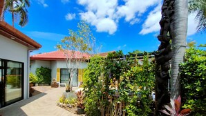Uma casa moderna com telhado de telhas vermelhas, varanda coberta e diversas plantas e árvores.
