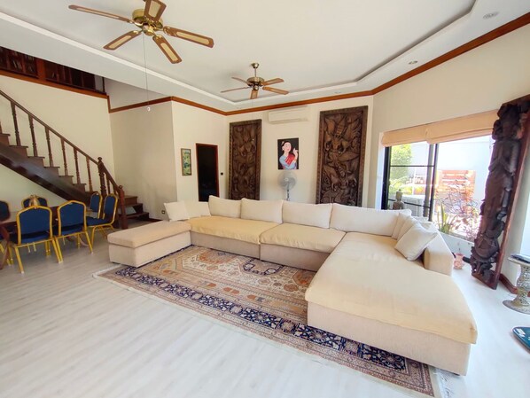 A spacious living room with a large L-shaped beige sofa, a patterned rug, a ceiling fan, and a staircase leading to an upper floor. - LUXURY VILLA WITH A VIEW OF TALAY, 400 M FROM THE BEACH - PATTAYA VACATION HOME (Muang Pattaya)