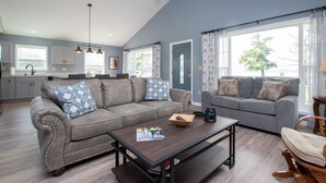 A modern living room with a grey sofa, a coffee table, and a wooden chair. There is a kitchen in the background with white cabinets and a window. - Seneca Spirit: Gorgeous Views of Seneca Lake! (Geneva)