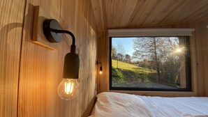 Un intérieur en bois comprenant un lit, un luminaire fixé au mur et une fenêtre donnant sur des arbres et des collines.