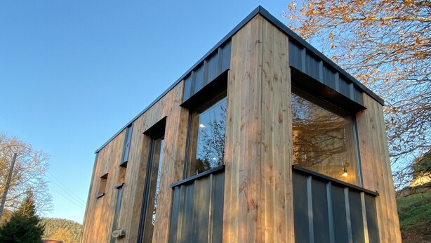 Une cabane en bois moderne, dotée de grandes fenêtres, d’un toit en pente et d’une pile de bois de chauffage à sa base.