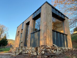 A modern wooden cabin with large windows, a sloped roof, and a stack of firewood at its base. - Homnest "Désirée" - Tiny House Vosgienne (3 pers.) (Saint-Dié-des-Vosges)