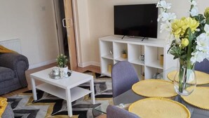 A modern living room with a glass coffee table, grey and yellow patterned rug, a sofa, a TV on a white stand, and a vase with flowers.