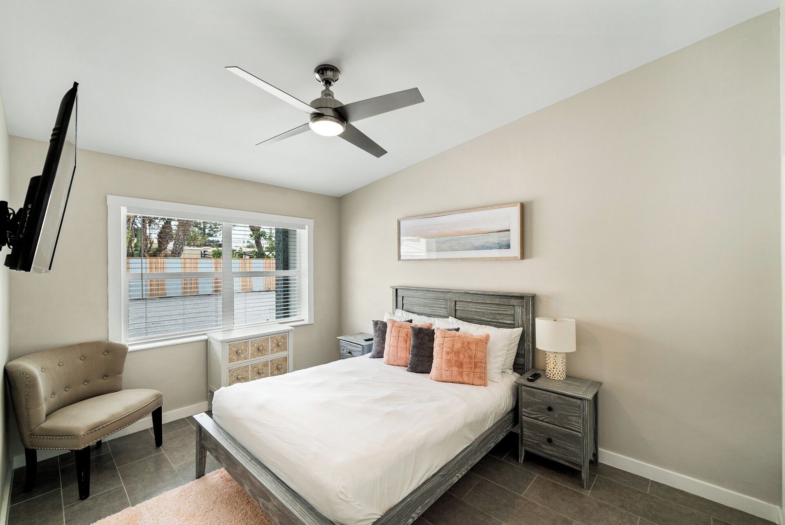 A bedroom with a bed, bedside tables, a chair, a ceiling fan, a window with blinds, and a wall-mounted artwork.