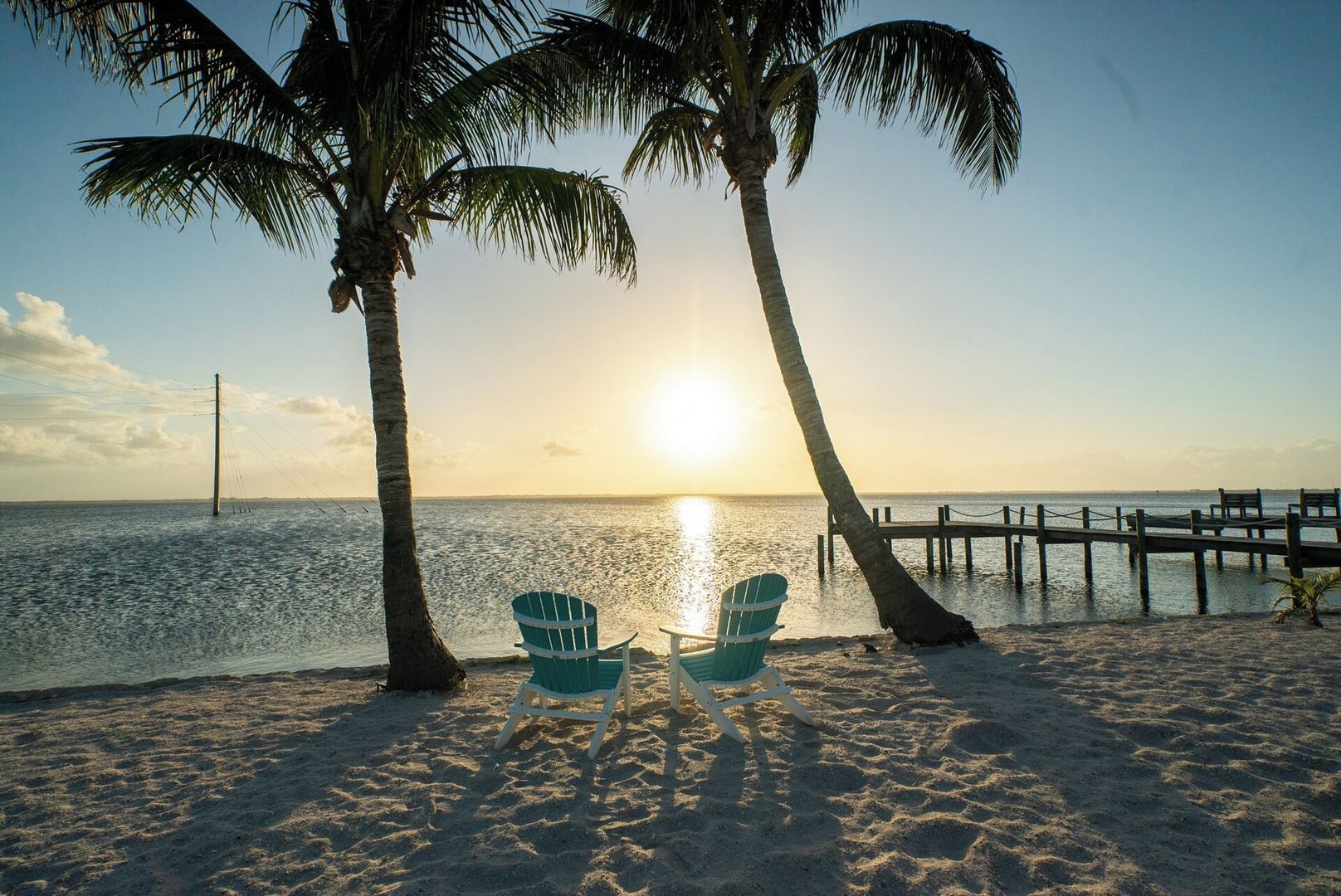 A beach scene with two chairs facing the ocean, palm trees, and a wooden pier extending into the water.