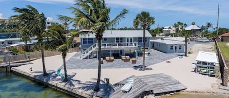 Una casa moderna con un muelle que se adentra en una masa de agua tranquila, rodeada de palmeras y un cielo despejado.