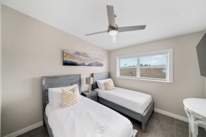 A hotel room with two beds, a ceiling fan, a window with blinds, and a painting on the wall.