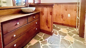 A bathroom with a wooden vanity, stone flooring, a window with blinds, and a wall-mounted light fixture.