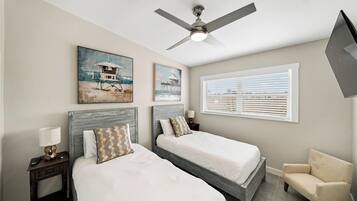 A hotel room with two beds, a ceiling fan, artwork on the walls, a window with blinds, and a chair.