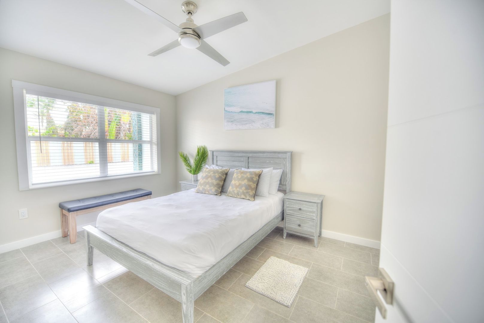 Une chambre à coucher comprenant un lit, des tables de chevet, un banc, un ventilateur de plafond, une fenêtre avec des stores et un tableau accroché au mur.