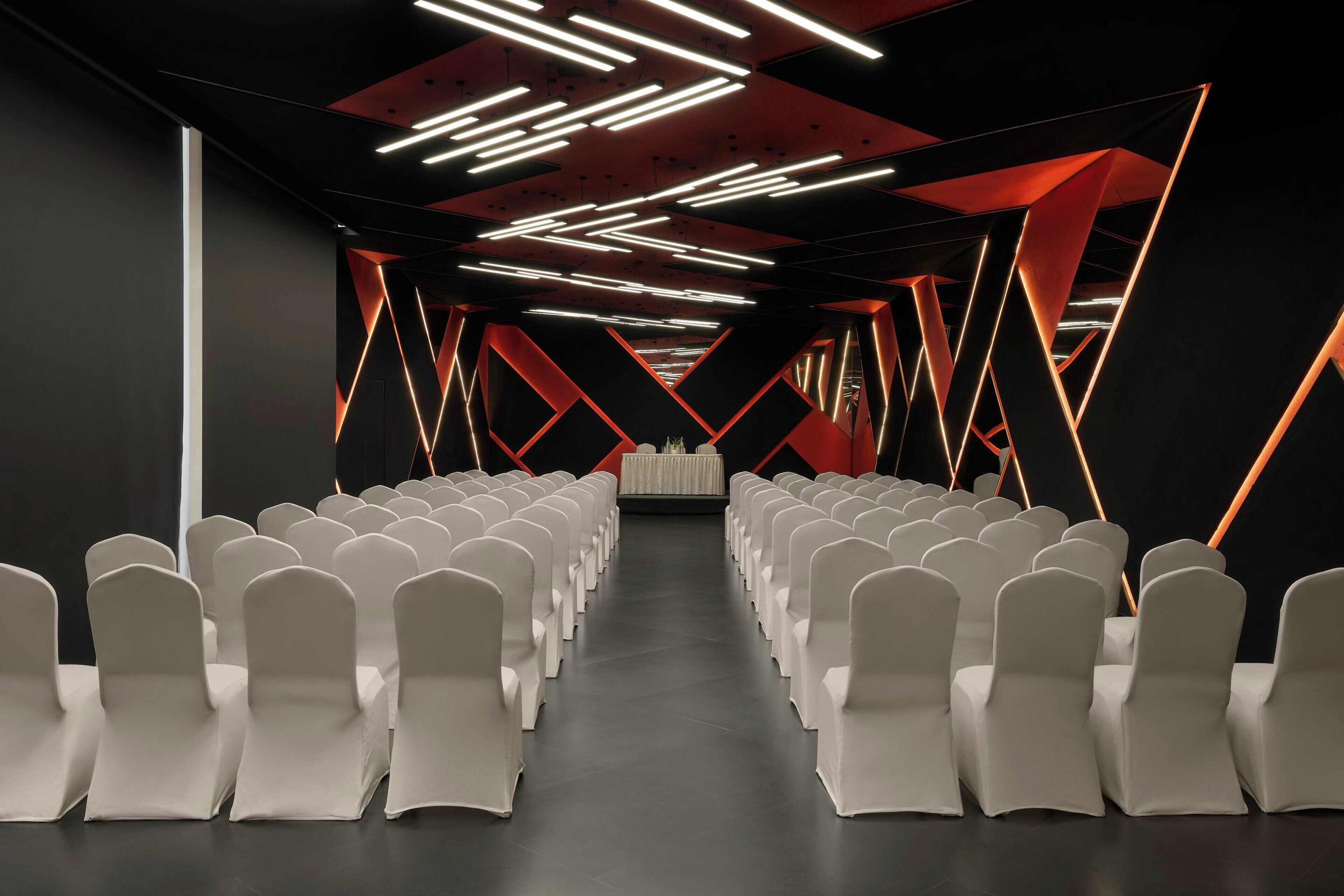A conference room with rows of white chairs, a stage at the front, and geometric ceiling lighting.