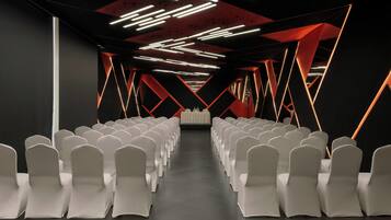 A conference room with rows of white chairs, a stage at the front, and geometric ceiling lighting.