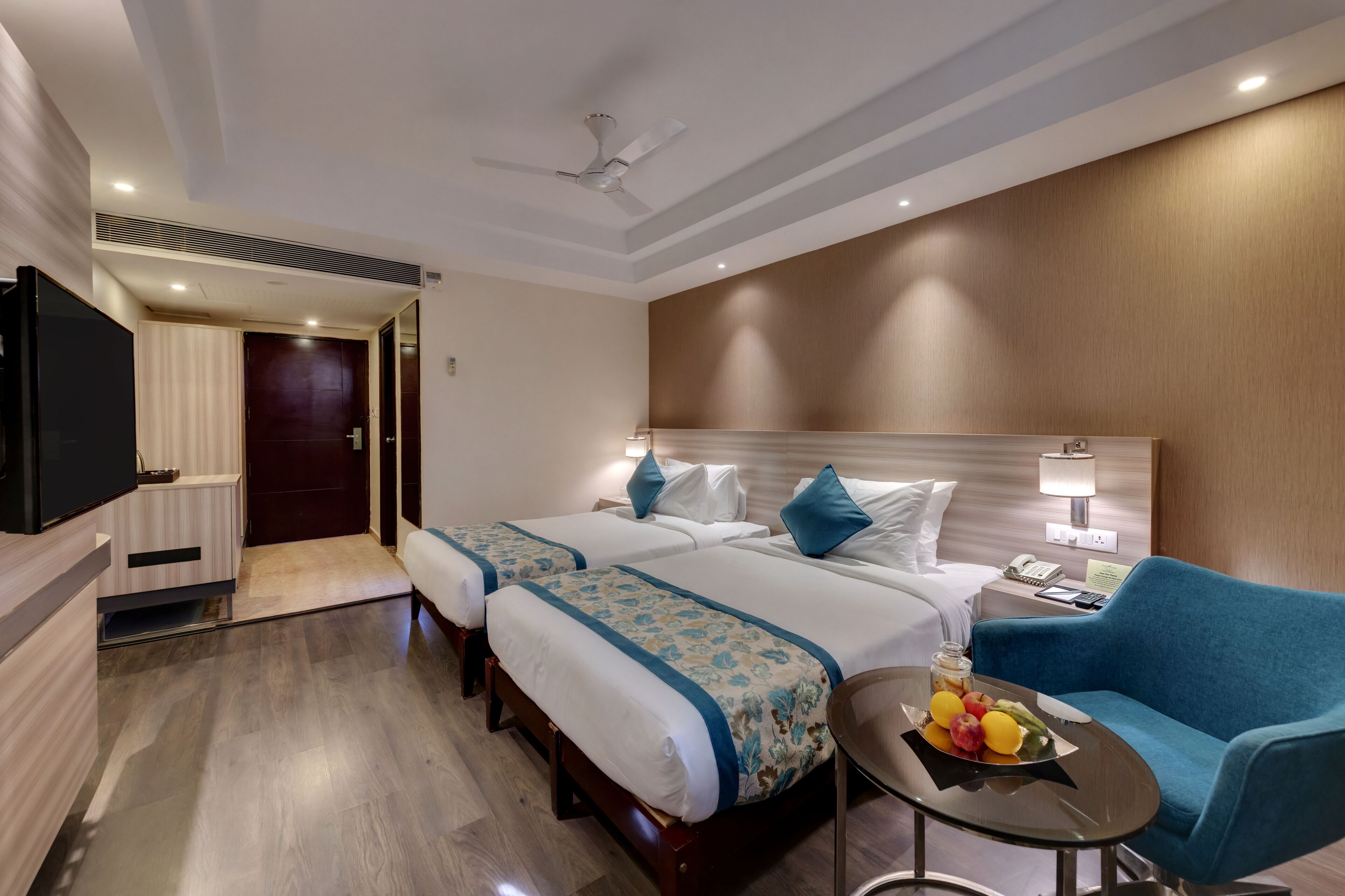 A hotel room with two beds, a TV, a desk with a lamp, and a coffee table with fruit.