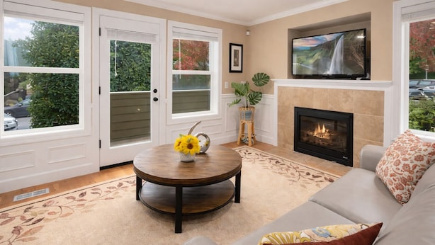 A living room with a fireplace, a sofa, a round coffee table with a vase of flowers, and a television mounted above the fireplace. - Perch - Cozy Ensuites: 3BR/3BA townhome with parking (Seattle)