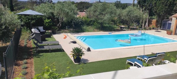 A rectangular swimming pool with a clear blue water surface, surrounded by a concrete deck. There are lounge chairs and a small table on the deck, and a potted plant near the pool's edge.