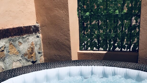 A hot tub with a textured edge, surrounded by a stone wall and a metal fence with greenery behind it.