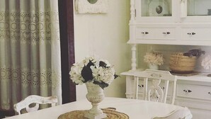 A dining area with a white table, chairs, and a vase of flowers. There is a white cabinet with glass doors and a mirror on the wall.