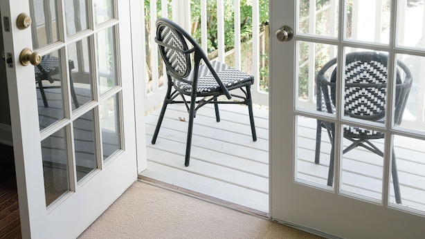 Vue depuis une pièce dont la porte est blanche et qui donne sur un balcon agrémenté de deux fauteuils en osier et d’une balustrade blanche.