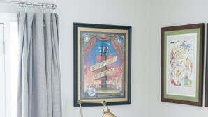 Une pièce avec un fauteuil blanc, un lampadaire, des rideaux gris et deux affiches encadrées accrochées au mur.