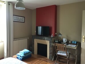 A hotel room with a bed, a desk with a chair, a television on a stand, a fireplace, and a radiator. - Beautiful stone house with a swimming pool in a secluded natural setting (Pondaurat)