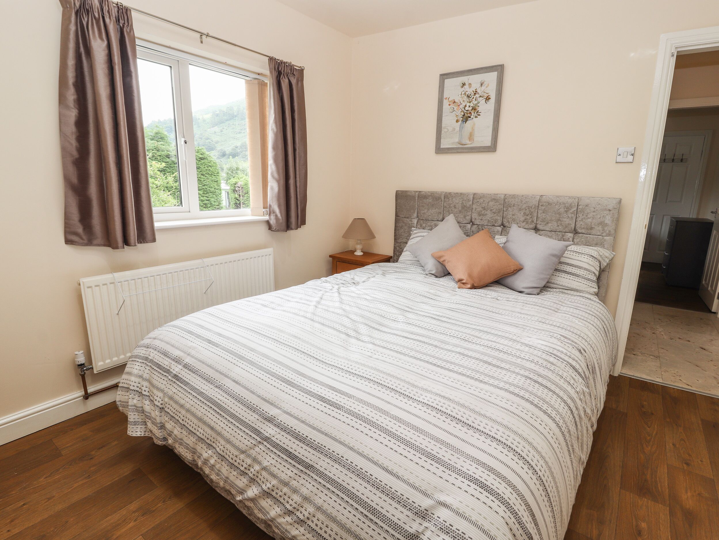 A bedroom with a bed, a window with curtains, a radiator, a bedside table with a lamp, and a framed picture on the wall.