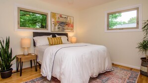 A bedroom with a large bed, two bedside lamps, a nightstand, a potted plant, and two windows with views of trees. - Warm & light-filled home with gym (Ann Arbor)