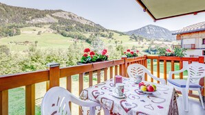 A balcony with a table set for two, a wooden railing, and a view of a green valley with mountains in the background. - Le cœur des Evettes - One Bedroom Apartment, Sleeps 4 (Flumet)