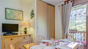A dining area with a wooden table set for four, a television on a cabinet, and a scenic view through a window. - Le cœur des Evettes - One Bedroom Apartment, Sleeps 4 (Flumet)