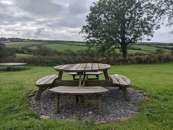 Una mesa de picnic de madera con bancos en un campo cubierto de hierba, rodeada de árboles y un paisaje rural.
