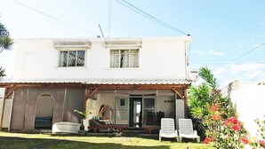 A two-story building with a flat roof, a porch with a wooden railing, and a satellite dish on the roof. There are two windows on the upper floor and a door on the ground floor.