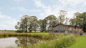 A modern lodge with a stone foundation and large windows, situated beside a calm pond and lush greenery. - Henry Haven (Preston)