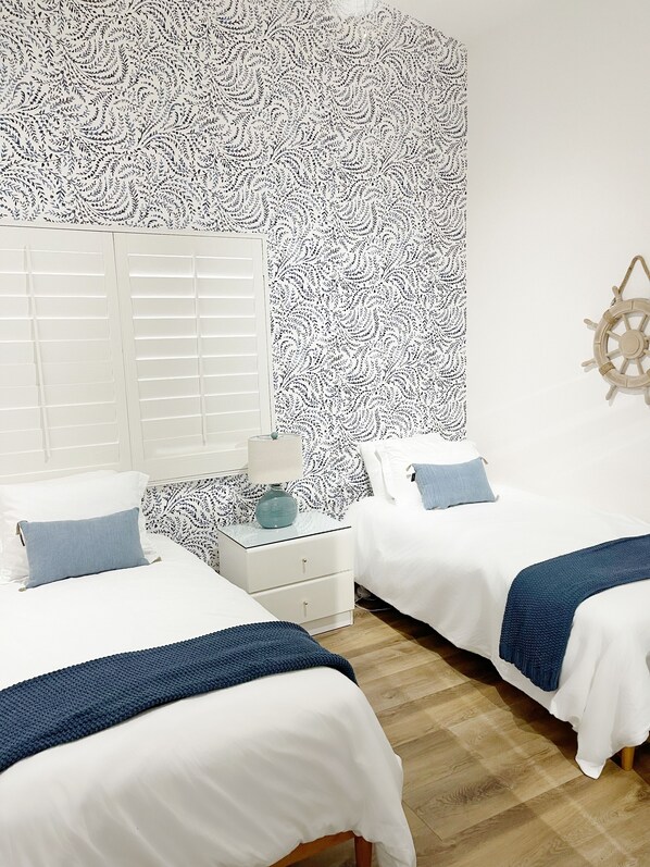 A bedroom with two beds, white bedding, a patterned wall, a wooden floor, and a ship's wheel decoration.