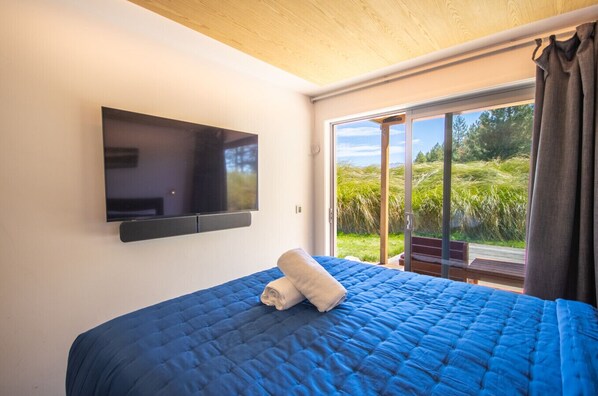 A bedroom with a large bed, a mounted TV, and a sliding glass door leading to a balcony with a view of greenery. - Clever Cube - Amazing Alpine Studio Unit (Twizel)