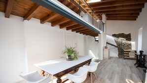 Un comedor amplio con una larga mesa de madera, sillas blancas y una planta en maceta. En el fondo, se ve una escalera con un muro de piedra y barandilla de metal.