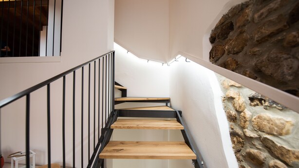 Una escalera con escalones de madera y barandillas negras que conduce a una habitación con pared de piedra y ventana con vistas.