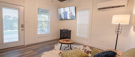 A spacious living room with a ceiling fan, a flat-screen TV mounted on the wall, and a large window with blinds.