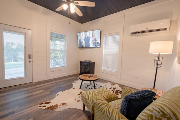 A spacious living room with a ceiling fan, a flat-screen TV mounted on the wall, and a large window with blinds. - Charming farmhouse, full kitchen, private back deck, fire pit, quiet area (Ringold)