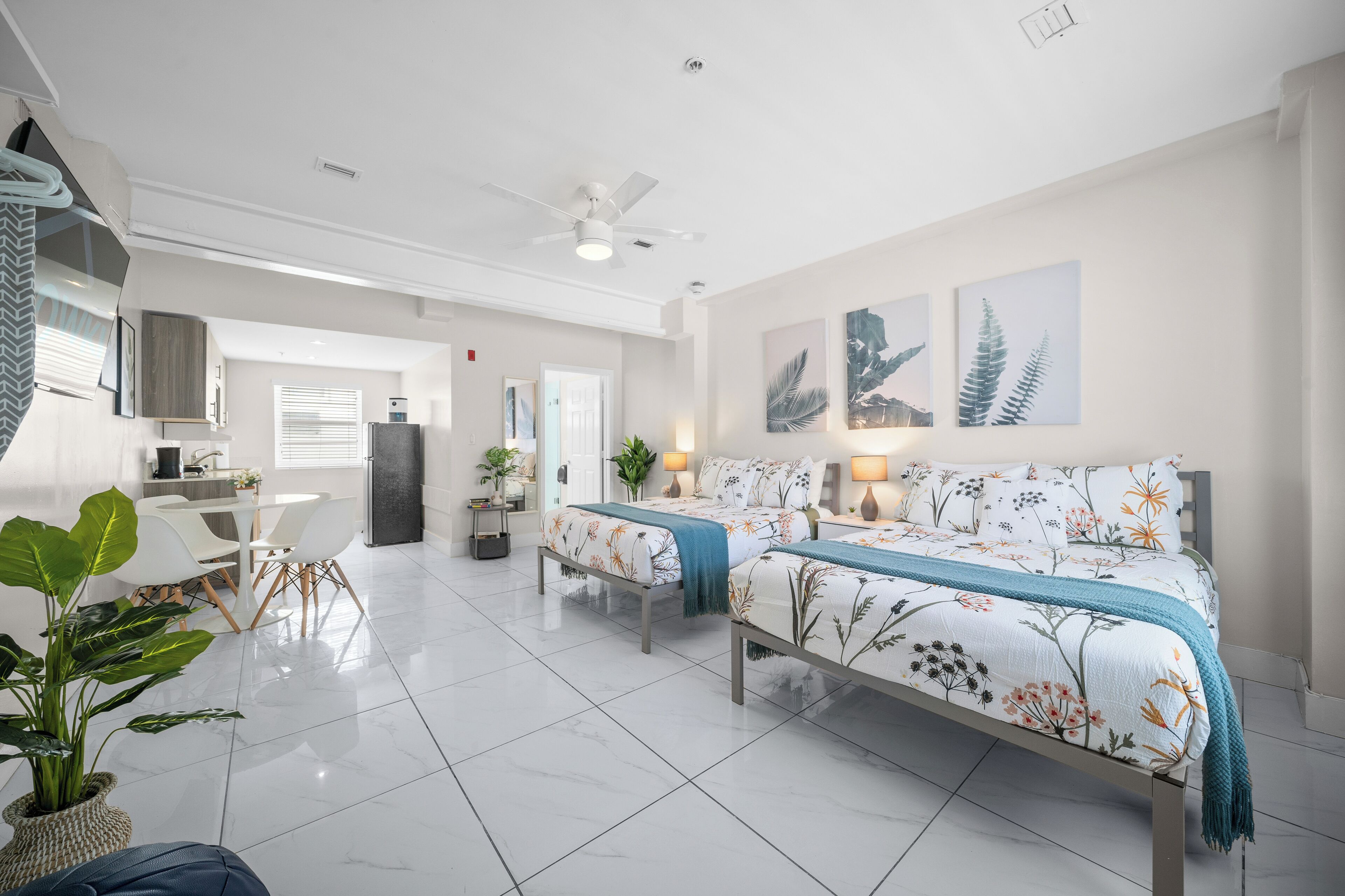 A modern hotel room with two beds, a dining area, and a kitchenette.