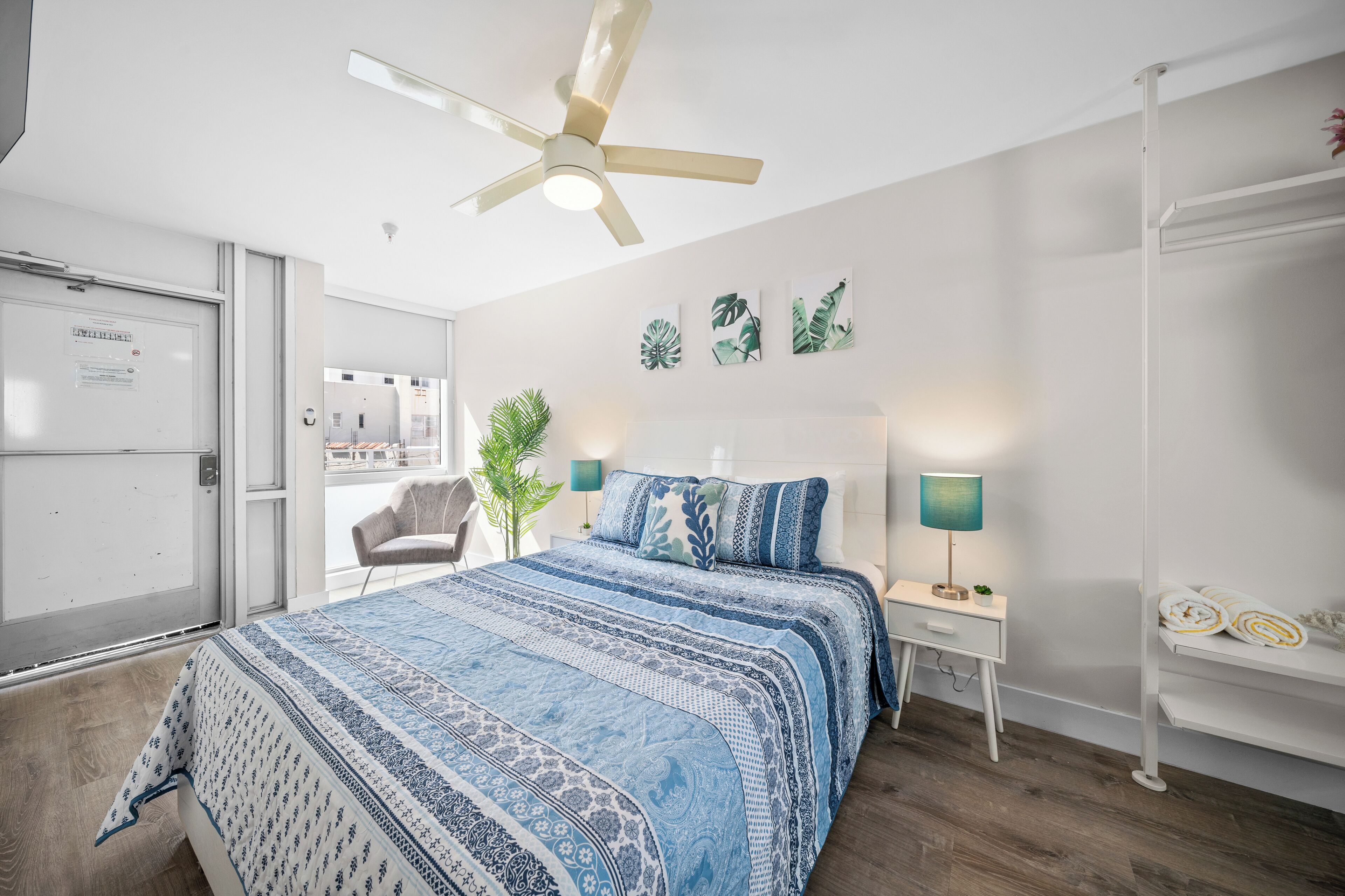 A bedroom with a bed, a ceiling fan, a nightstand, a lamp, a plant, and a view of the outside.