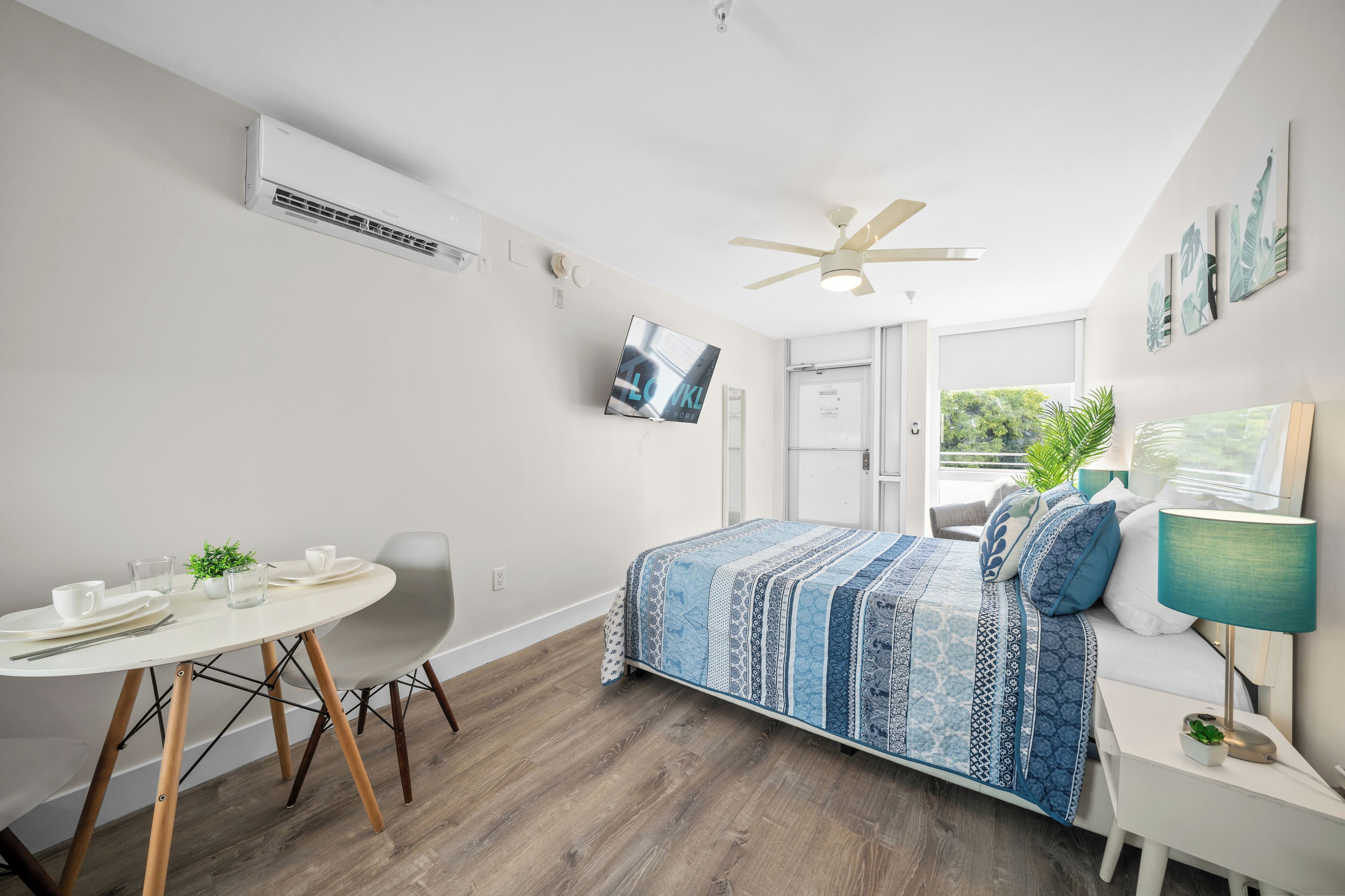A modern bedroom with a bed, a desk, and a chair. There is a ceiling fan, a TV, and a window with a view of greenery.