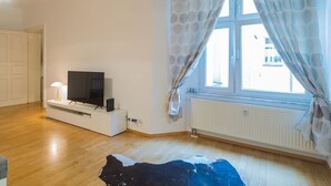 A room with a TV on a white stand, a radiator, and a window with curtains. - fully furnished 2-room apartment in Berlin (Berlin)