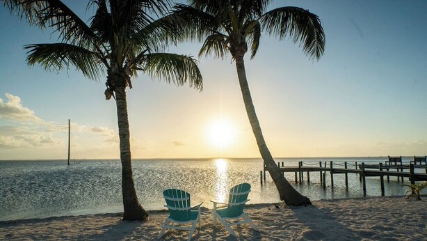 A beach scene with two chairs facing the ocean, palm trees, and a wooden pier extending into the water. - Chasing Pelicans #9 - Waterfront - Sunset Views (Cocoa Beach)