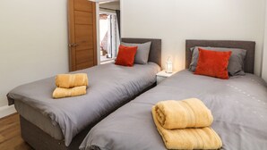 A double room with two beds, each with grey bedding and orange pillows. There is a wooden door and a small white nightstand with a lamp between the beds.