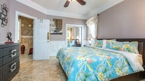 Une chambre à coucher comprenant un lit, un ventilateur de plafond, une commode et une porte donnant sur une autre pièce.