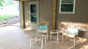 Un porche abrité avec une terrasse en bois, deux fauteuils en osier, une petite table ronde et une porte avec une fenêtre.