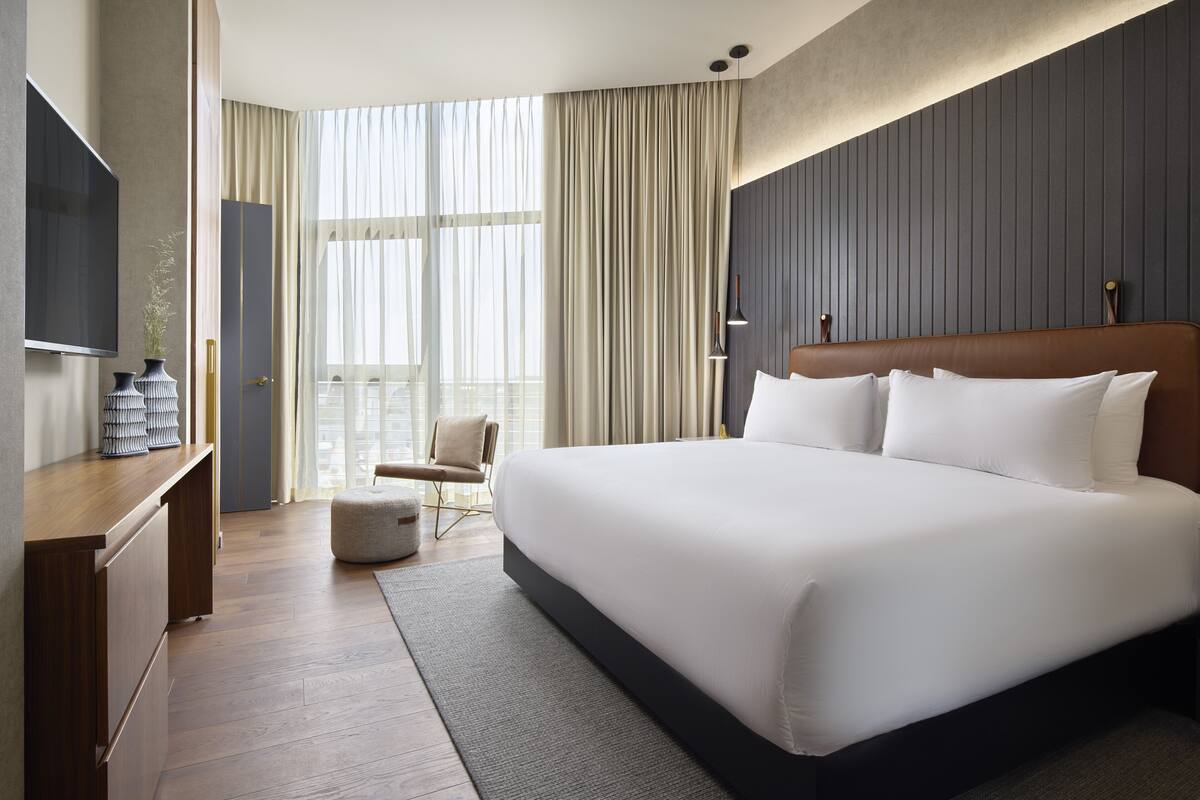 A modern hotel room with a large bed, a flat-screen TV, a desk, and a view of the outdoors.