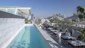 Piscina en la azotea con sillones de descanso y plantas en macetas, con vistas a un paisaje urbano.