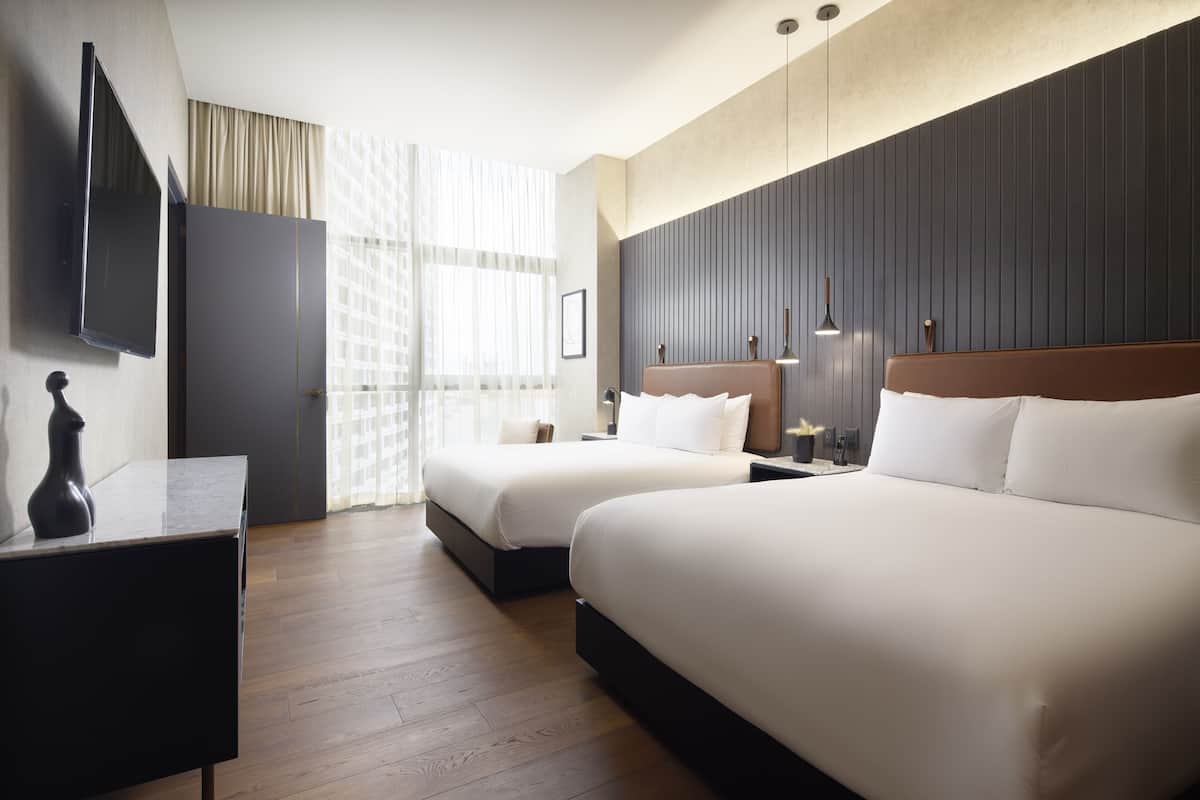 A hotel room with two beds, a flat-screen TV, a desk, and a large window with curtains.